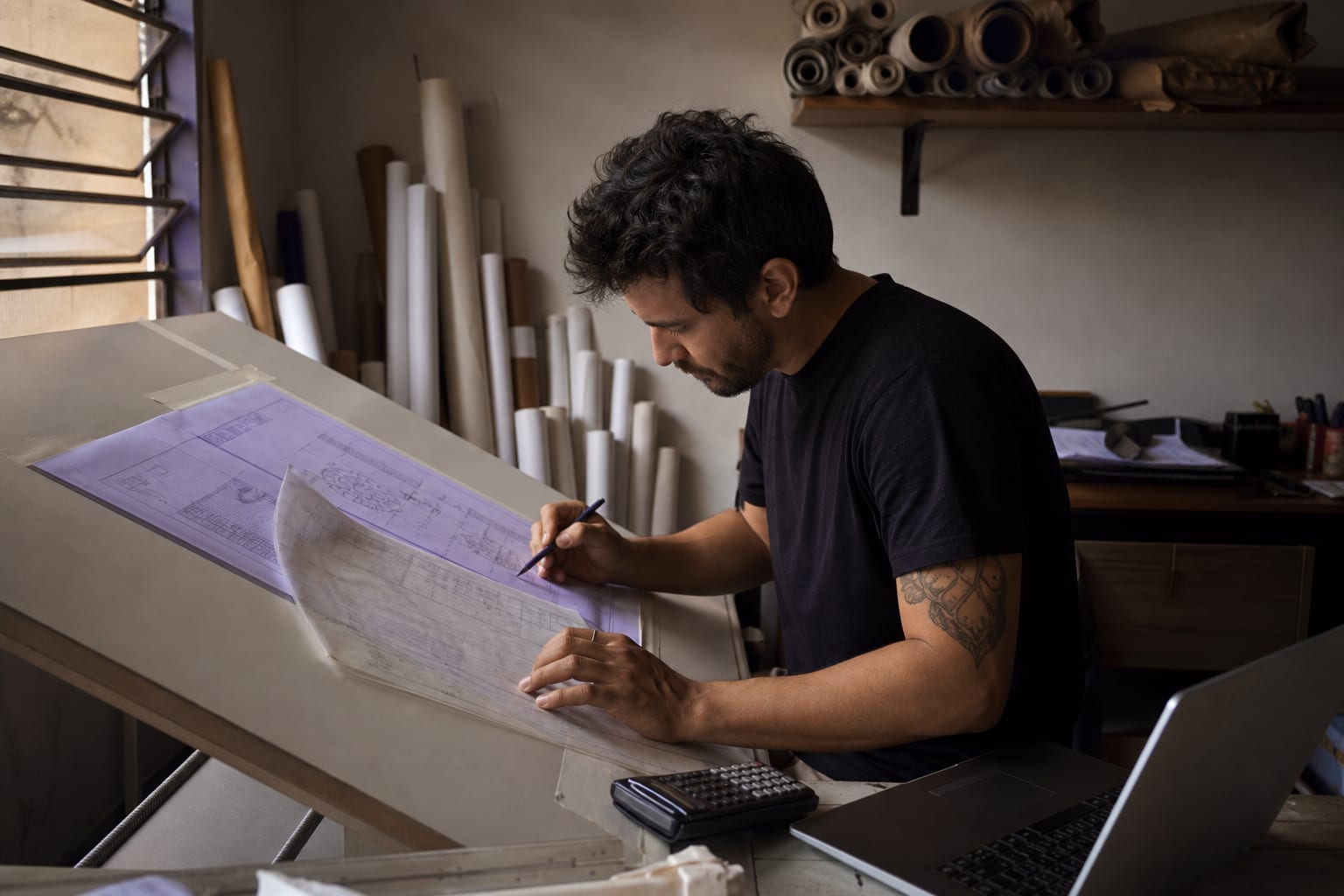 Arquiteto trabalhando em orçamento com planta impressa, escalímetro e notebook sobre a mesa do escritório
