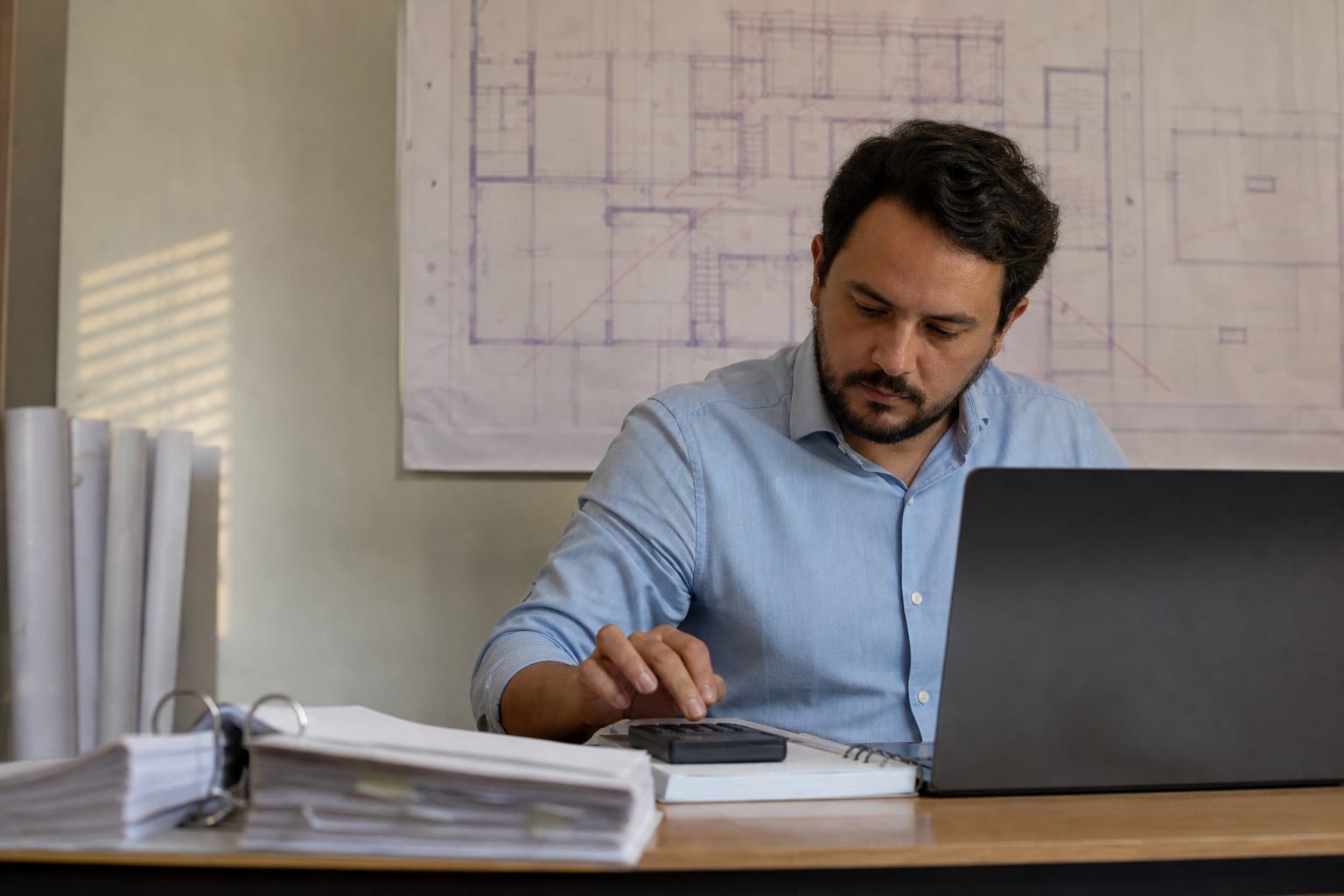 Sócio de construtora conferindo números em calculadora de mesa ao lado do notebook no escritório